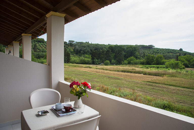 Covered balcony with table and chairs overlooking fields and wooded hills in Perivoli, Corfu